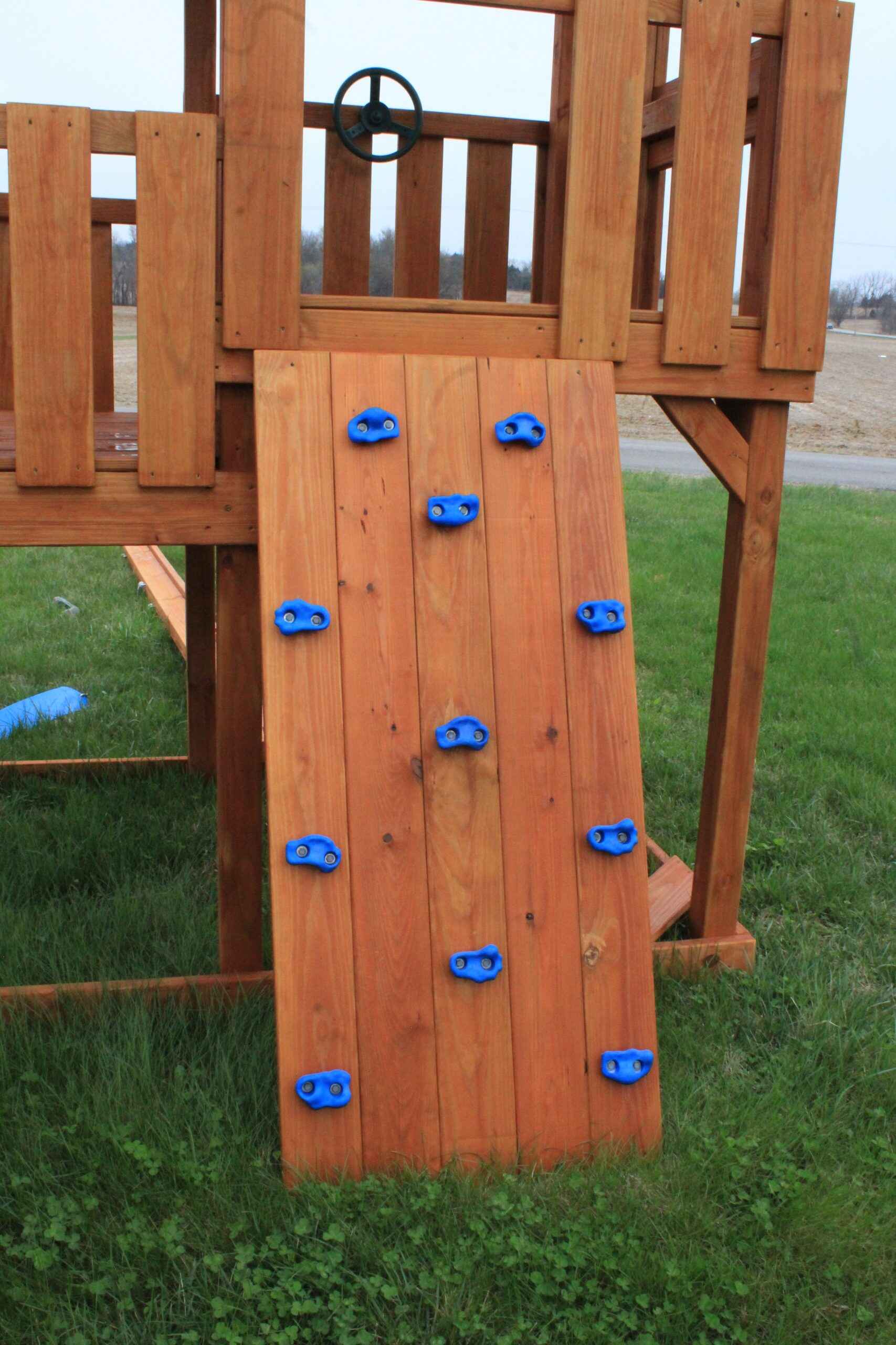 Features multi-colored rock holds mounted on an angled climbing surface. The holds can be repositioned to create different climbing challenges, and the wall includes safety ropes for added security.